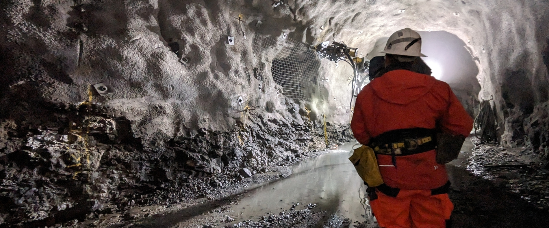 A person in orange protective clothing stands in a lit rock tunnel with water puddles, rock anchors, and a drilling machine in the background.