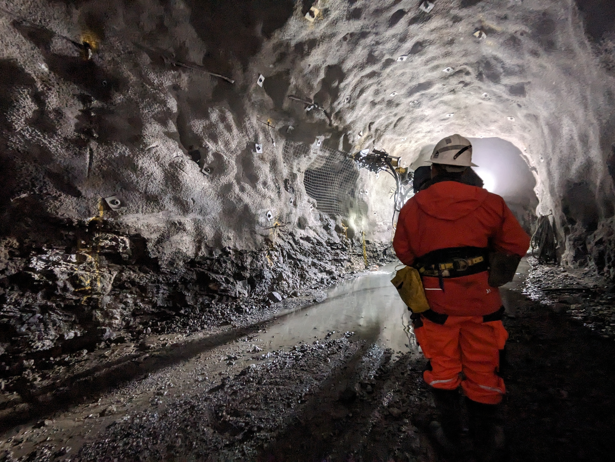 A person in orange protective clothing stands in a lit rock tunnel with water puddles, rock anchors, and a drilling machine in the background.